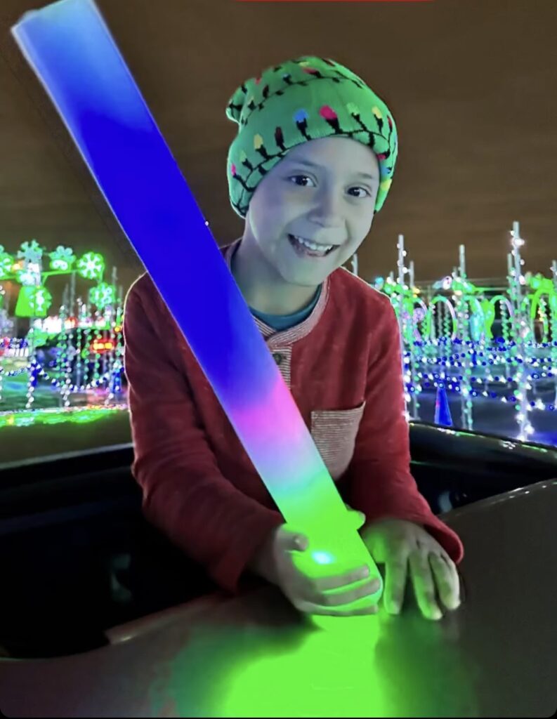 A boy pops out of the sunroof of the car holding onto a large glow stick and wearing a light up hat with Christmas lights on it.